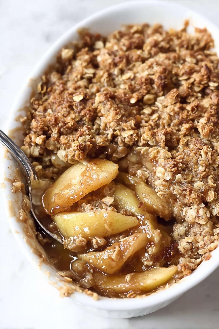 The dish is shown in a white oval baking dish filled with a warm baked apple crumble. The top layer is a coarse golden brown crumb made up of oats, flour, and sugar, with a rough and crunchy texture. Beneath this topping, tender apple slices coated in a glossy, syrupy caramel sauce fill the middle layer, showing a soft texture with a light amber color. The apples have slightly browned edges from baking. A silver spoon is pressed into the crumble on the right side, slightly lifting some of the crumb and apple layers. The dish rests on a white marbled surface. Photo taken with an iphone --ar 2:3 --v 7