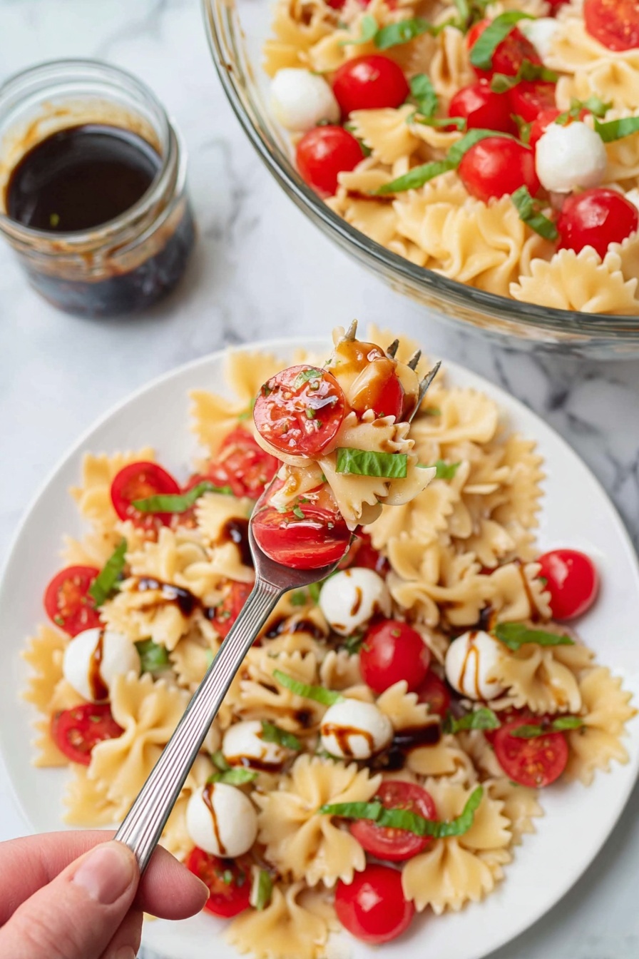 The image shows a white plate filled with farfalle pasta mixed with bright red cherry tomatoes, small white mozzarella balls, and thin strips of green basil scattered on top. There is a light drizzle of dark balsamic glaze over the pasta. A spoon holds a serving of the pasta mixture close to the camera, showing the pasta’s bowtie shape and the fresh tomato halves and whole mozzarella balls in clear detail. In the background, a glass bowl with more pasta and tomatoes sits on a white marbled surface, along with a small glass jar of dark balsamic sauce and a spoon inside it. Photo taken with an iphone --ar 2:3 --v 7