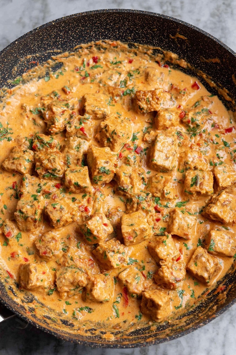 Creamy Sun-Dried Tomato Cashew Tofu - The image shows a pan filled with a creamy sauce that has a light orange color mixed with green herbs and small red bits. In the sauce, there are many evenly sized, light brown cubes that are partly covered by the sauce. The pan is round with a dark, patterned surface, and it sits on a white marbled background. The texture of the sauce looks thick and smooth with visible small pieces blending in photo taken with an iphone --ar 2:3 --v 7