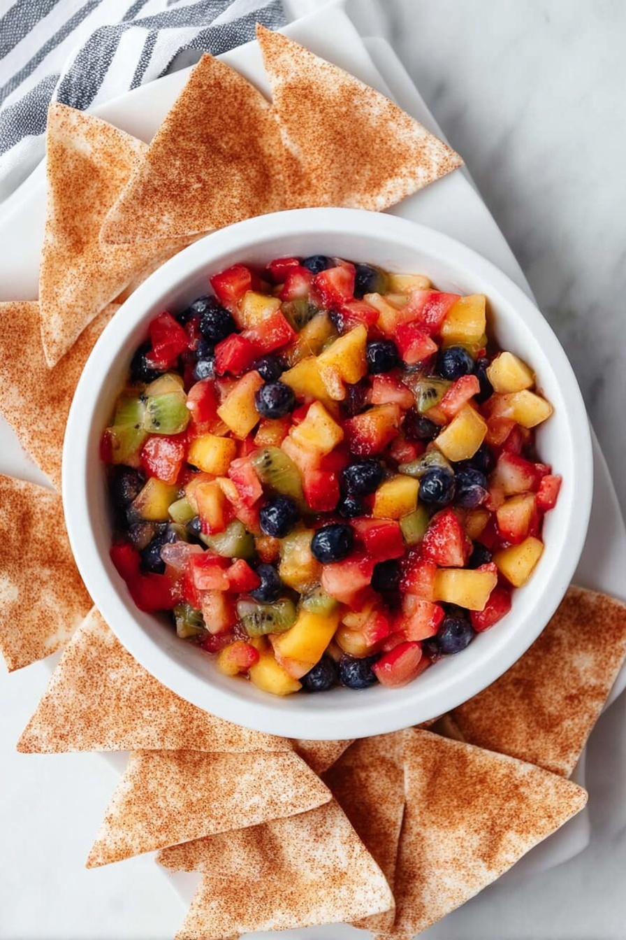 A white bowl filled with colorful fruit salsa sits in the center of a white plate, surrounded by several triangular cinnamon-dusted chips. The salsa is made of small, uneven pieces of red strawberries, deep blue blueberries, yellow mango, and pale green kiwi mixed together, creating a bright, textured surface with a glossy finish. The chips have a light brown tone with a visible layer of cinnamon spice, arranged loosely around the bowl. The entire scene is set on a white marbled surface with a striped cloth partially visible in the corner. Photo taken with an iphone --ar 2:3 --v 7