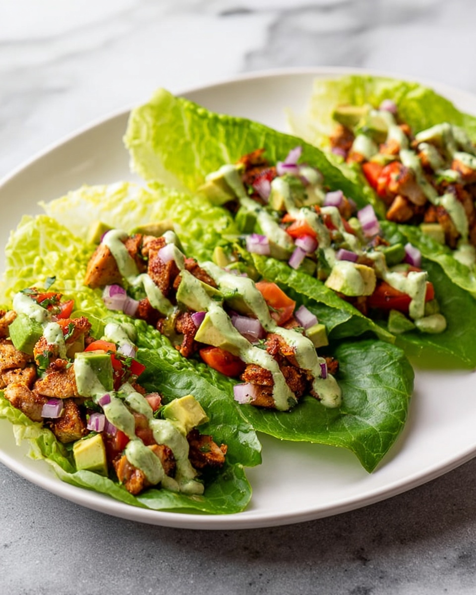 The image shows three lettuce wraps placed on a white plate over a white marbled surface. Each wrap starts with a bright green romaine lettuce leaf, forming a natural bowl shape. Inside the leaves are layers of cooked, slightly browned chicken pieces spread evenly. On top of the chicken, there are small chunks of fresh avocado in a light green color and diced red tomatoes scattered around. Small bits of purple onion are also sprinkled throughout. A light green creamy sauce is drizzled generously over the fillings in each wrap. In the background, there is a wedge of lime partially visible. photo taken with an iphone --ar 2:3 --v 7