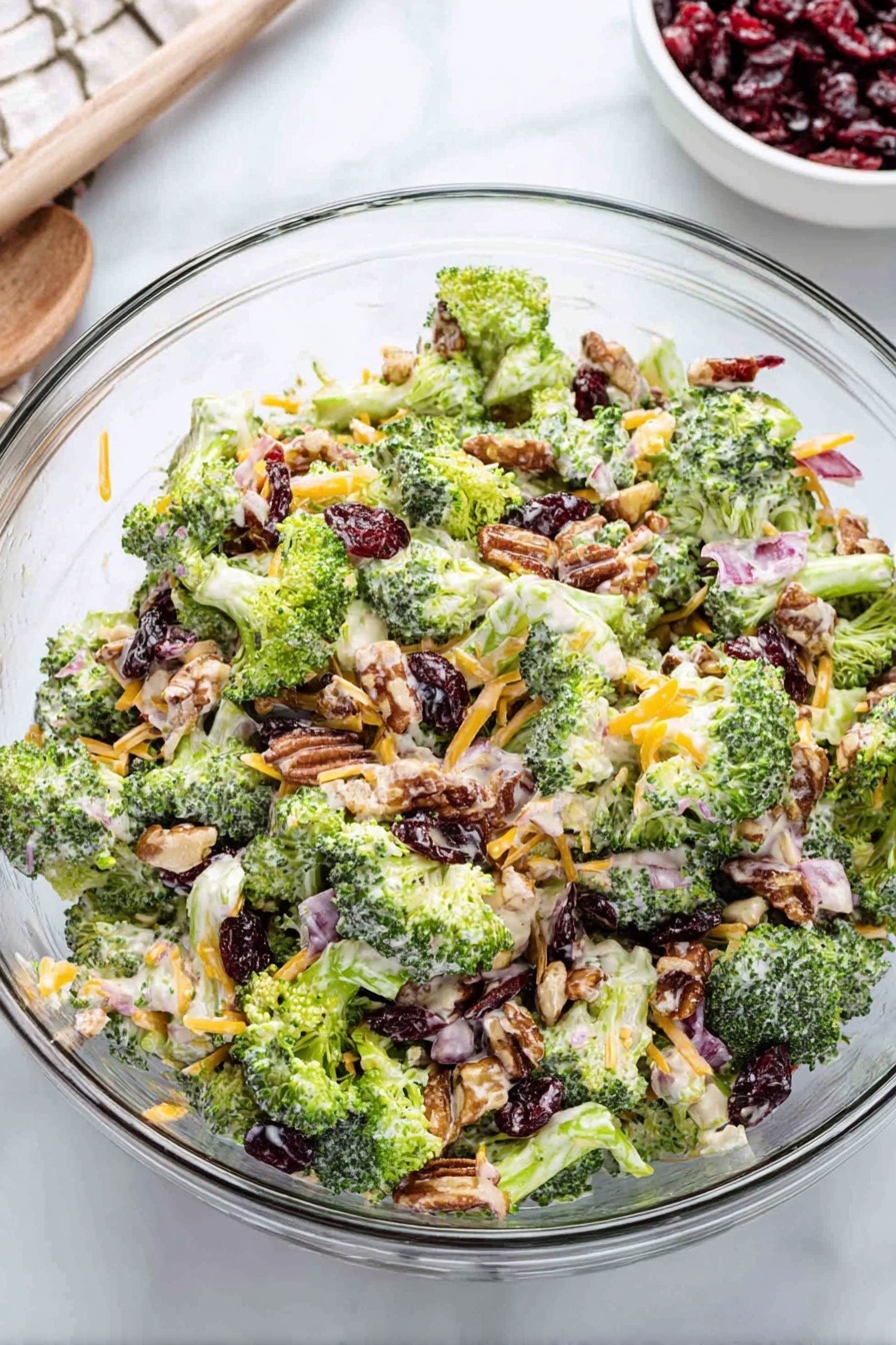 A clear glass bowl is filled with a colorful broccoli salad consisting of bright green broccoli pieces evenly mixed with shredded orange cheese, dark red dried cranberries, small light brown nut pieces, and bits of pale purple onion. The salad is lightly coated with a creamy white dressing, giving it a slightly glossy texture. The bowl sits on a white marbled surface with a small white bowl of extra dried cranberries partially visible in the background. Photo taken with an iphone --ar 2:3 --v 7