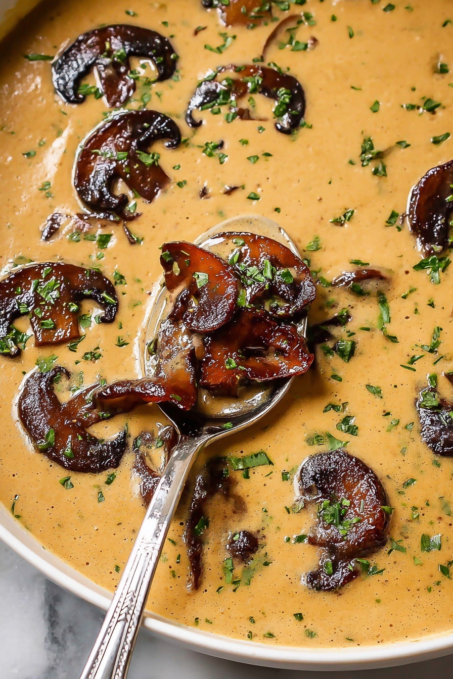 The image shows a creamy light brown sauce base with a smooth texture filling the frame. On top of this sauce, there are several dark brown, caramelized mushroom slices scattered around, some partially submerged in the sauce. Bright green chopped herbs, likely parsley or chives, are sprinkled evenly over both the sauce and mushrooms, adding a fresh contrast in color. A shiny silver spoon dips into the sauce from the lower right side, lifting a portion that includes multiple mushroom slices and herbs. The whole dish sits on a white marbled surface, enhancing the warm colors of the food. photo taken with an iphone --ar 2:3 --v 7
