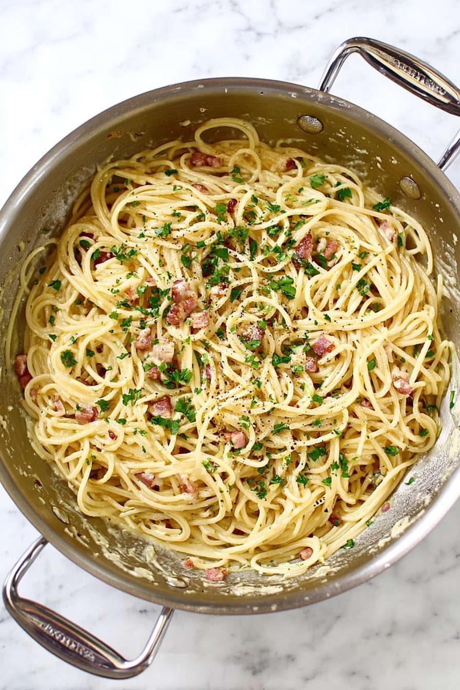 A shiny silver pan with two handles holds creamy spaghetti mixed with small pieces of cooked bacon and sprinkled with chopped green herbs and black pepper. The long pasta strands are coated evenly with a light yellow sauce, and the pan sits on a white marbled surface, showing some bits of sauce stuck to the pan edge. photo taken with an iphone --ar 2:3 --v 7