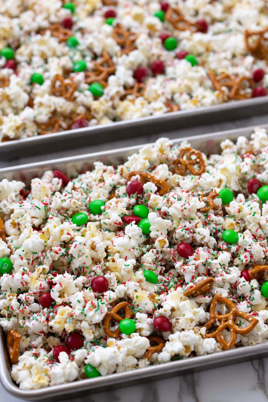 The image shows two metal trays filled with a festive snack mix spread out evenly. The mix has three main layers: the first is white popcorn scattered all over with a fluffy texture; mixed throughout are small, round red and green candies adding a bright, shiny pop of color; the third layer consists of small light brown pretzel twists scattered evenly, giving a crunchy texture. There are also tiny red and green sprinkles sprinkled all over the popcorn mix, creating a festive and colorful look. The trays are placed on a white marbled surface. photo taken with an iphone --ar 2:3 --v 7
