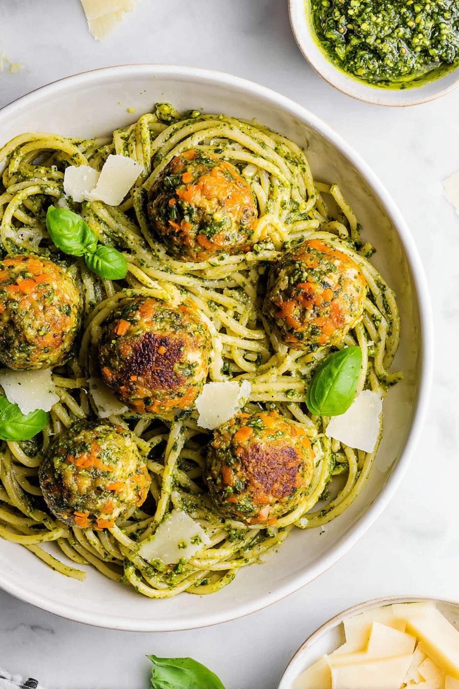 A white bowl filled with long, thick spaghetti noodles coated in bright green pesto sauce forms the base layer. On top are six round meatballs that have a golden brown, slightly crispy texture with bits of green herbs and orange carrot pieces visible. Scattered around the meatballs and noodles are small fresh green basil leaves and a few thin, pale yellow cheese shavings. The bowl sits on a white marbled surface, with a small white dish of chunky green pesto and a white plate of pale cheese shards visible at the bottom right. photo taken with an iphone --ar 2:3 --v 7