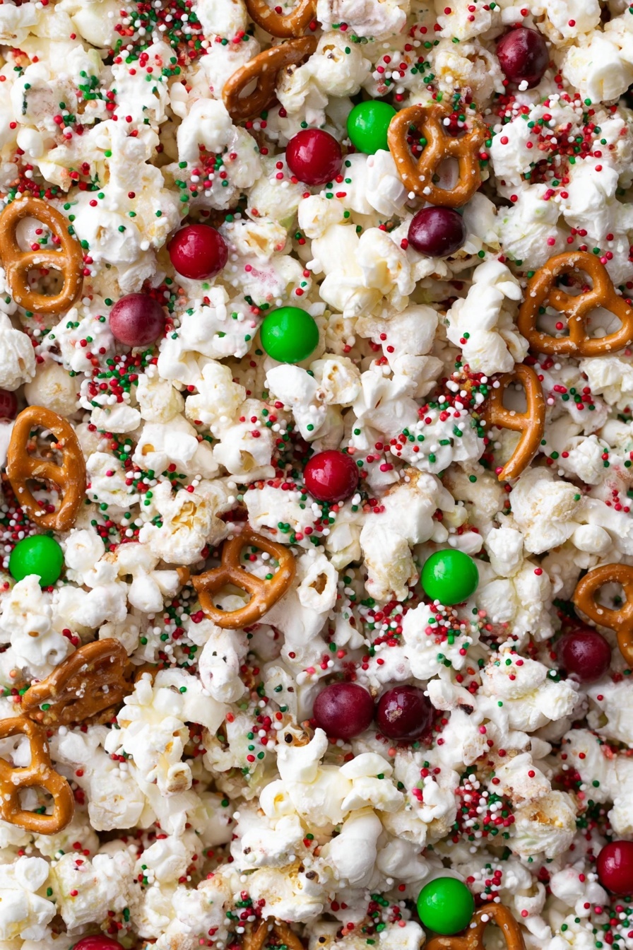The image shows a close-up view of a mixed snack with three main layers. The base layer consists of white popcorn clusters that have a light, fluffy texture. Scattered evenly throughout are small, round red and green candy pieces that add bright spots of color. Interspersed within are golden-brown pretzels with a firm, twisted shape. The entire mix is sprinkled with tiny round sprinkles in red, green, and white, creating a festive look. The background is a white marbled texture. photo taken with an iphone --ar 2:3 --v 7