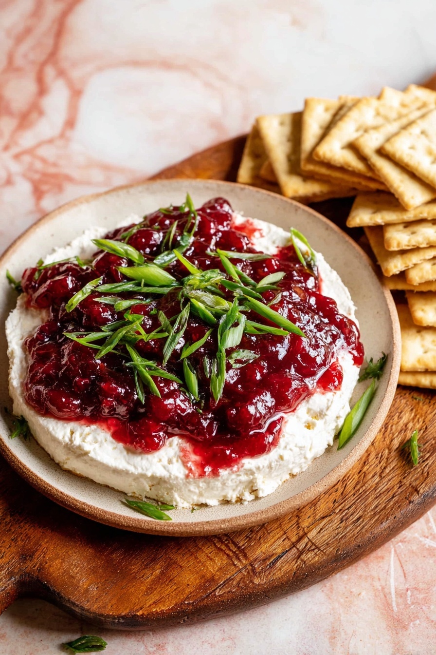 A round white plate with a creamy white cheese layer spread thickly on the bottom, topped with a thick, glossy dark red layer of cranberry sauce spread evenly over it. On top, bright green chopped scallions are scattered, adding contrast. The plate sits on a brown wooden cutting board, alongside a pile of square beige crackers stacked on the right. The background features a white marbled surface with soft pink tones. Photo taken with an iphone --ar 2:3 --v 7