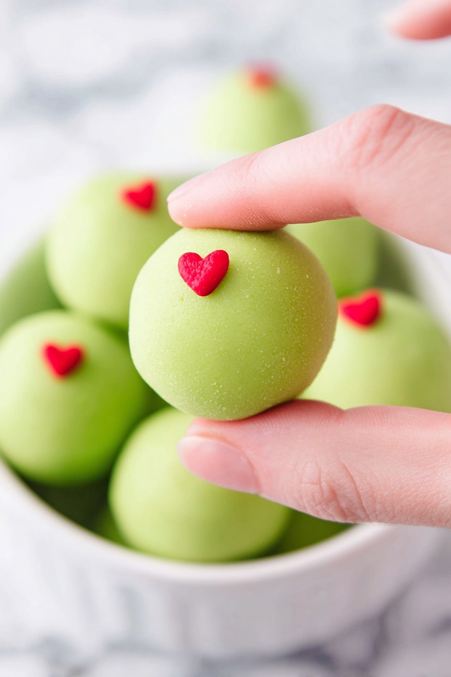 A close-up shows a woman's hand holding a smooth, round green ball with a small red heart decoration on top. Below, a white bowl is filled with more green balls, each topped with a tiny red heart. The background is soft and white marbled, giving a clean and bright look. The green balls have a smooth texture and look slightly matte, with the red hearts adding a pop of bright color. photo taken with an iphone --ar 2:3 --v 7
