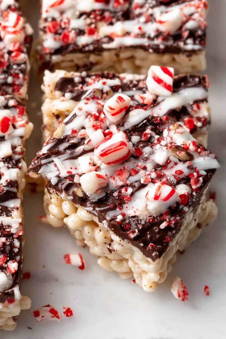 The image shows several square pieces of a crispy rice treat stacked on a white marbled surface. Each piece has three main layers: the bottom and middle layers are light golden and crispy, made of puffed rice cereal mixed with a sticky, smooth marshmallow layer, while the top layer is a thin coating of dark chocolate drizzled unevenly, sprinkled with crushed red and white peppermint candy bits. Two whole peppermint candies sit near the front right of the frame, with some blurred stacks of the treats in the background, creating depth. The photo taken with an iphone --ar 2:3 --v 7