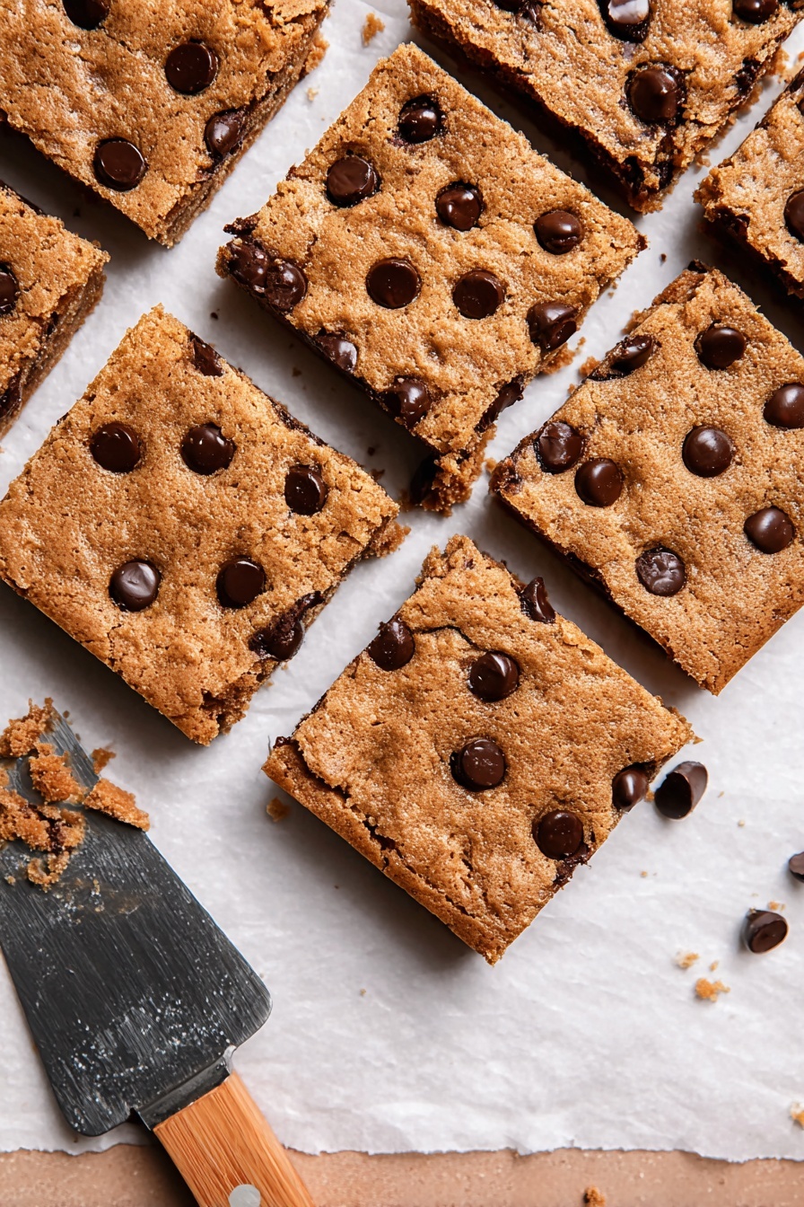 The image shows nine square chocolate chip cookie bars arranged closely on a white paper-lined surface. Each cookie bar is golden brown with a slightly rough, cracked texture and scattered with shiny dark chocolate chips both on top and partially melted within. The bars look thick and soft, with some small crumbs around them. A white marbled surface is partly visible at the bottom with a spatula that has a wooden handle placed near the cookies. photo taken with an iphone --ar 2:3 --v 7