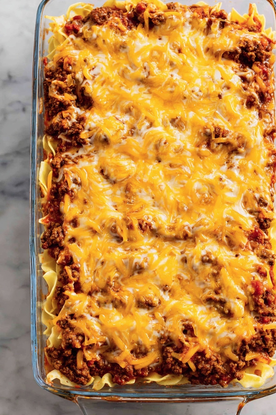 A clear glass rectangular dish filled with three layers: the bottom layer is pale yellow cooked pasta, the middle layer contains dark brown cooked ground meat mixed with red tomato sauce, and the top layer is melted cheese in bright orange and white colors evenly spread across the dish. The dish is set on a white marbled surface. photo taken with an iphone --ar 2:3 --v 7