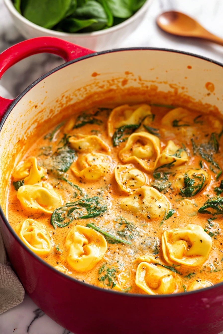 A close-up view of a red pot filled with creamy orange sauce mixed with yellow tortellini and green spinach leaves. The sauce looks thick and rich, coating the tortellini which float evenly throughout. The inside of the pot is white, showing some sauce splashes on the sides. In the blurred background, there is a white bowl with fresh green spinach and a wooden spoon resting next to the pot. The pot sits on a white marbled surface. photo taken with an iphone --ar 2:3 --v 7