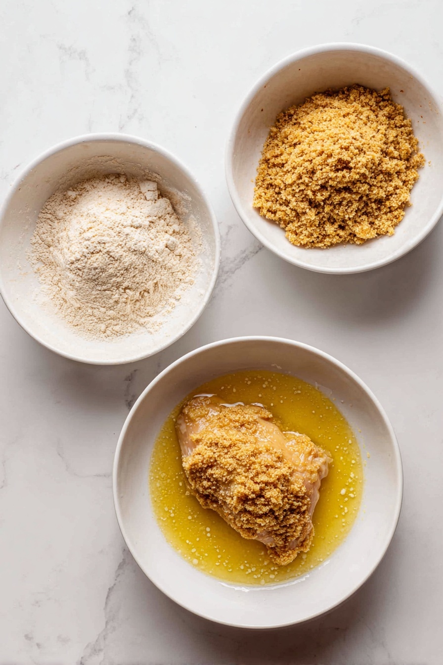 The image shows a white plate on a white marbled surface, holding two layers of golden-brown breaded chicken stacked on the right side. The top layer is sliced vertically to show the white, tender interior. On the left side of the plate, there is a serving of creamy mashed potatoes with a smooth, slightly lumpy texture, sprinkled lightly with chopped green herbs. Two lemon wedges are placed near the top and bottom edges of the plate, adding a bright yellow contrast. A gold fork rests near the bottom right of the plate, and some green herbs are scattered around the food. Photo taken with an iphone --ar 2:3 --v 7