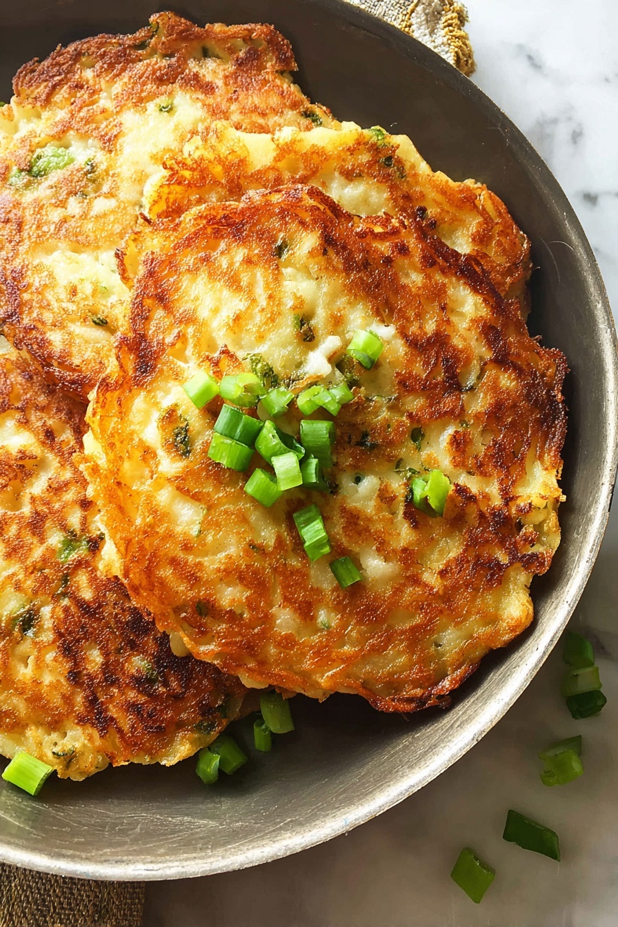 A white plate holds three golden brown potato pancakes stacked slightly overlapping, showing a crispy, textured surface with some rough edges. On top of the pancakes is a small dollop of white sour cream sprinkled with finely chopped green onions. More green onion pieces are scattered over the pancakes and the white marbled surface beneath. A fork and a spoon are partly visible at the plate's edge, resting on a dark blue cloth. Photo taken with an iphone --ar 2:3 --v 7