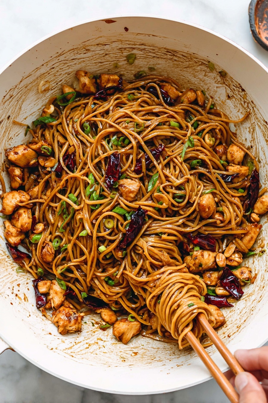 A large white wok filled with glossy brown noodles mixed with small, evenly cut golden-brown chicken pieces, scattered light brown peanuts, and dark red dried chili peppers. The noodles are slightly shiny from the sauce, and fresh green chopped spring onions are sprinkled throughout, adding bright green pops. A woman's hand is using wooden chopsticks to lift and twirl a bundle of noodles on the right side of the wok. The white marbled surface underneath highlights the colorful dish, and some sauce splashes are seen on the inside of the wok. Photo taken with an iphone --ar 2:3 --v 7