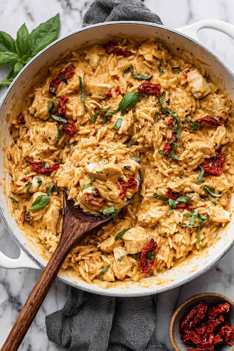 A white frying pan filled with a creamy orange sauce mixed with small pieces of chicken, orzo pasta, sun-dried red tomatoes, and fresh green basil leaves. The sauce coats all the ingredients evenly, with some visible black pepper sprinkled on top. A wooden spoon is scooping up a portion, showing the thick and rich texture of the dish. The pan sits on a gray cloth on a white marbled surface, with a small bowl of sun-dried tomatoes and a few loose basil leaves nearby. photo taken with an iphone --ar 2:3 --v 7